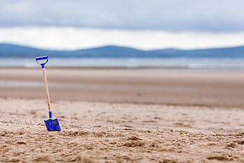 Ein Bild von einem Strand. Im Vordergrund steckt eine Spielschaufel im Sand. Im Hintergrund befindet sich das Meer und Berge.