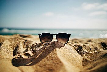 Ein Bild von einem Strand. Im Vordergrund steckt eine Sonnenbrille im Sand. Im Hintergrund befindet sich das Meer und blauer Himmel.