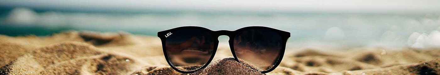 Ein Bild von einem Strand. Im Vordergrund steckt eine Sonnenbrille im Sand. Im Hintergrund befindet sich das Meer und blauer Himmel.